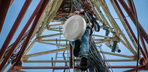 Telecommunications technician climbing a broadcast tower with safety harness and equipment, under a clear blue sky.